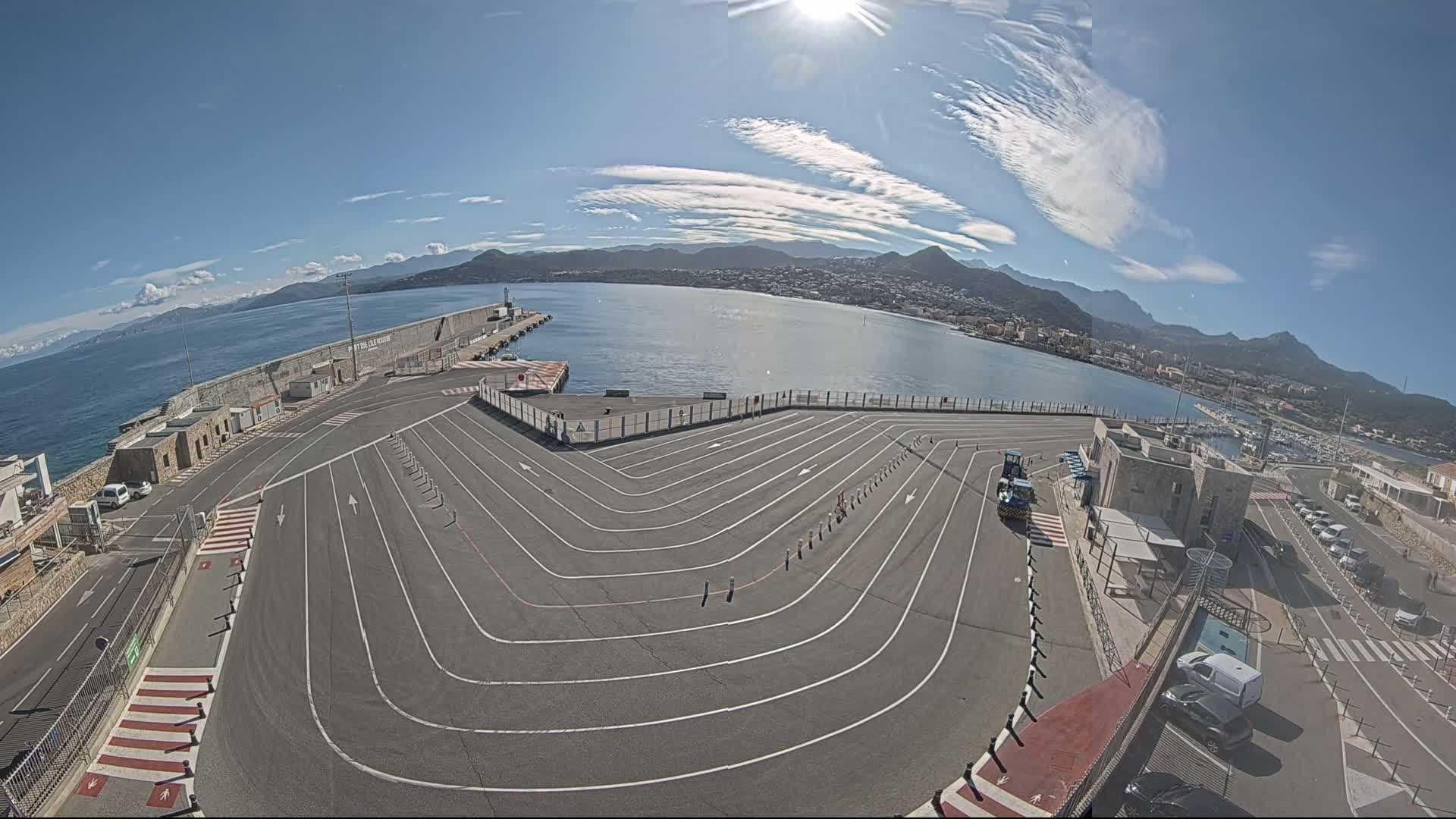 A large, paved area with marked lanes curves around a harbor, overlooking a calm sea and distant mountains under a mostly sunny sky.