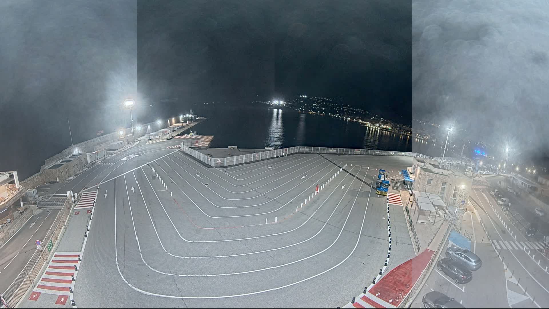 A nighttime aerial view of a ferry terminal with a large, curving vehicle staging area, under hazy conditions.