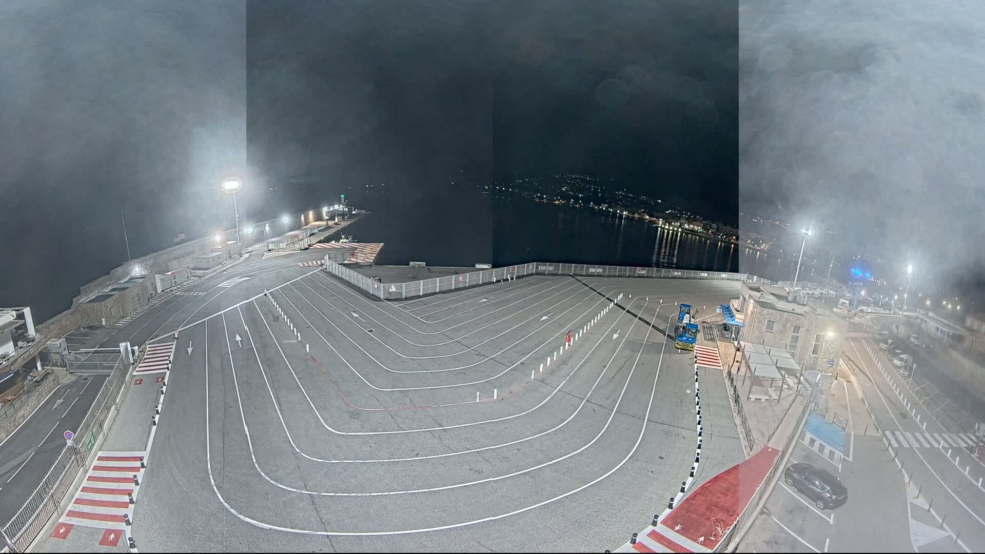 An empty, marked vehicle staging area at a port at night, under a hazy sky.