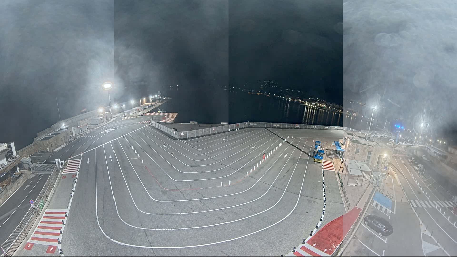 A nighttime, hazy view of a large, empty port area with white lane markings, a few vehicles, and a body of water in the background.