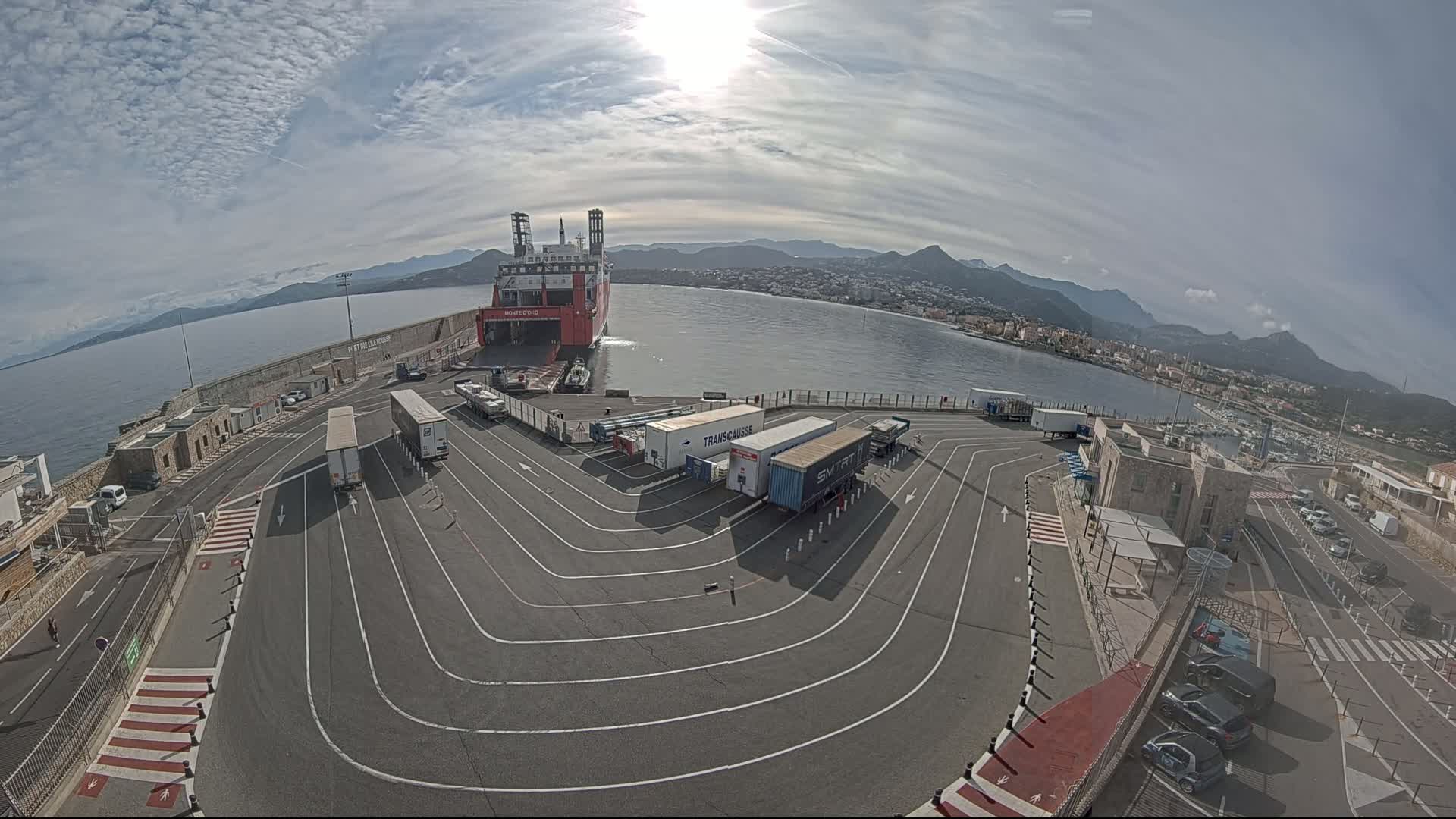 A large red ferry is docked at a port, with several semi-trailer trucks parked in a designated area, under a partly sunny sky.