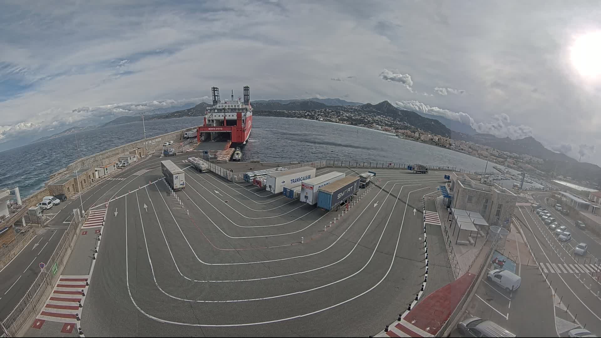 A ferry is docked at a port, with several trucks and cars parked nearby, under an overcast sky.