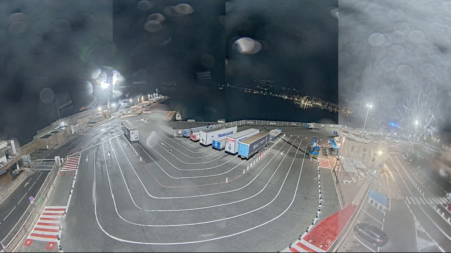 A nighttime view of a ferry terminal with several semi-trailer trucks parked in designated lanes, under rainy conditions.