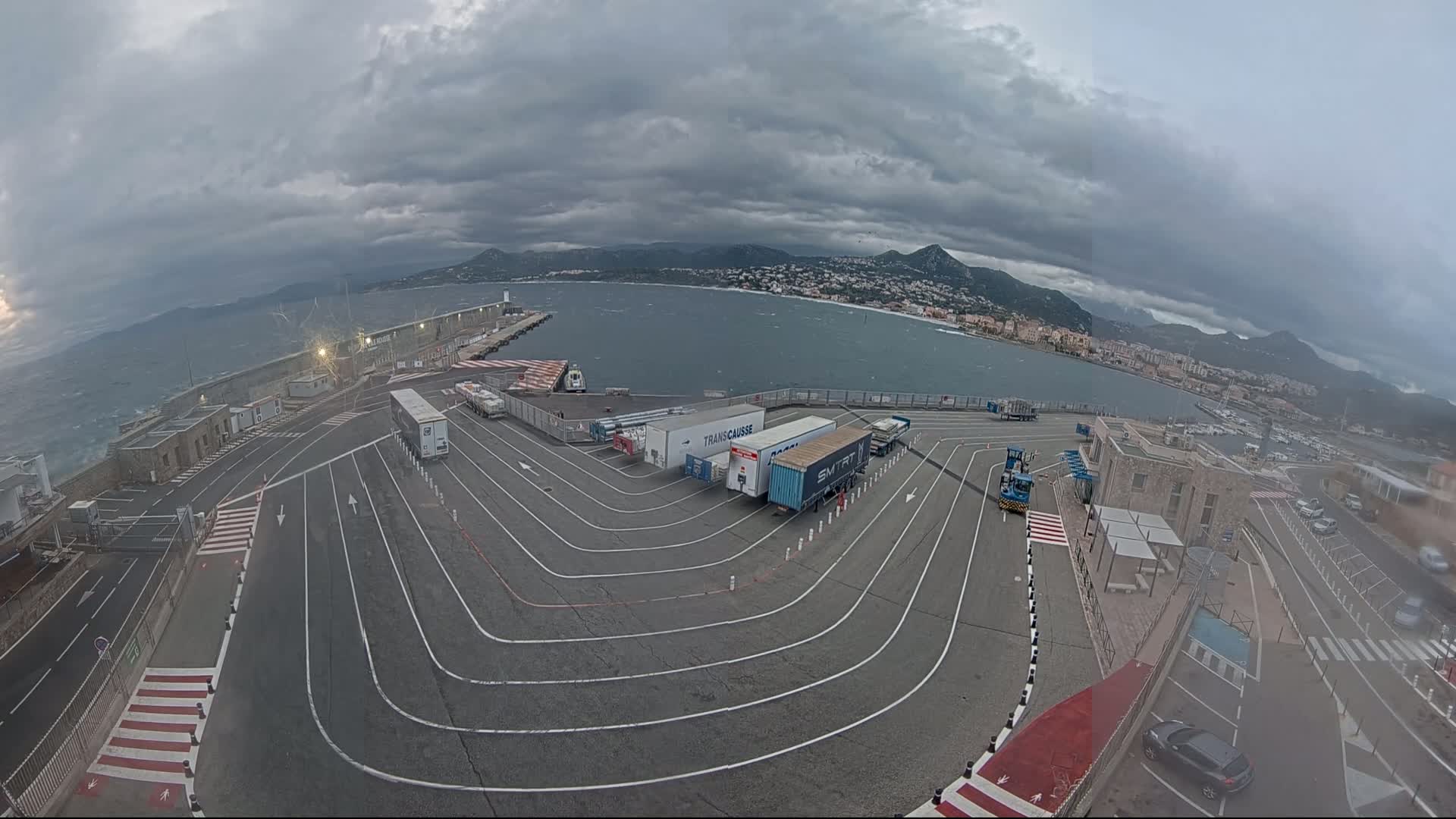 An aerial view shows a ferry terminal with several trucks parked in a designated area, overlooking a choppy sea under a cloudy sky and mountainous background.