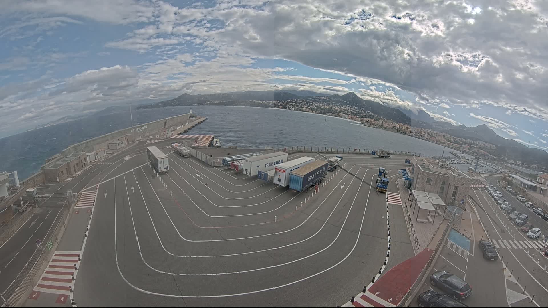An aerial view shows a port area with several semi-trailer trucks parked in designated lanes, a body of water with mountains in the background under a partly cloudy sky.