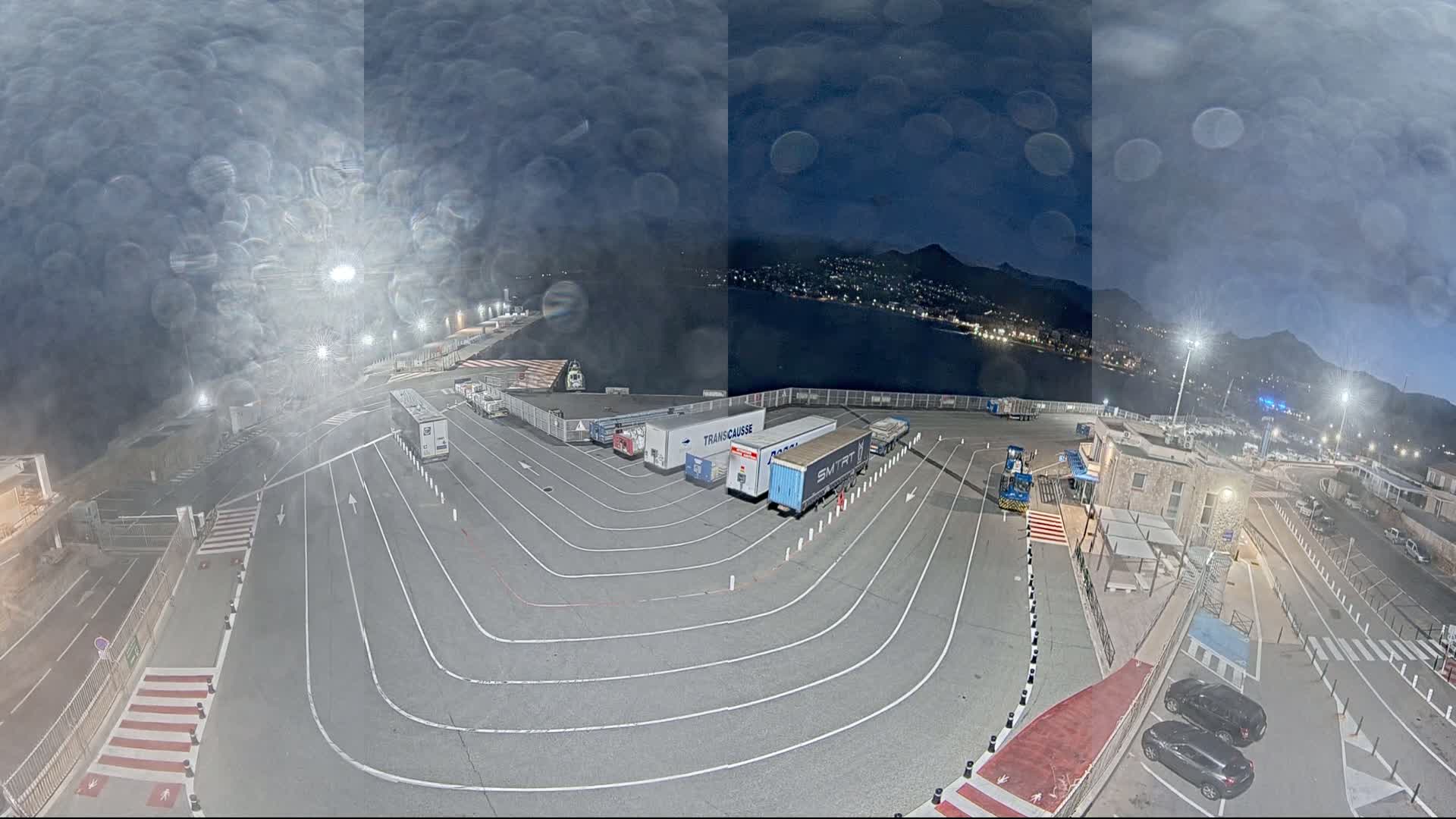 A nighttime, rain-streaked view of a port area with several trucks parked in designated lanes, overlooking a body of water and a distant city lit up against dark mountains.