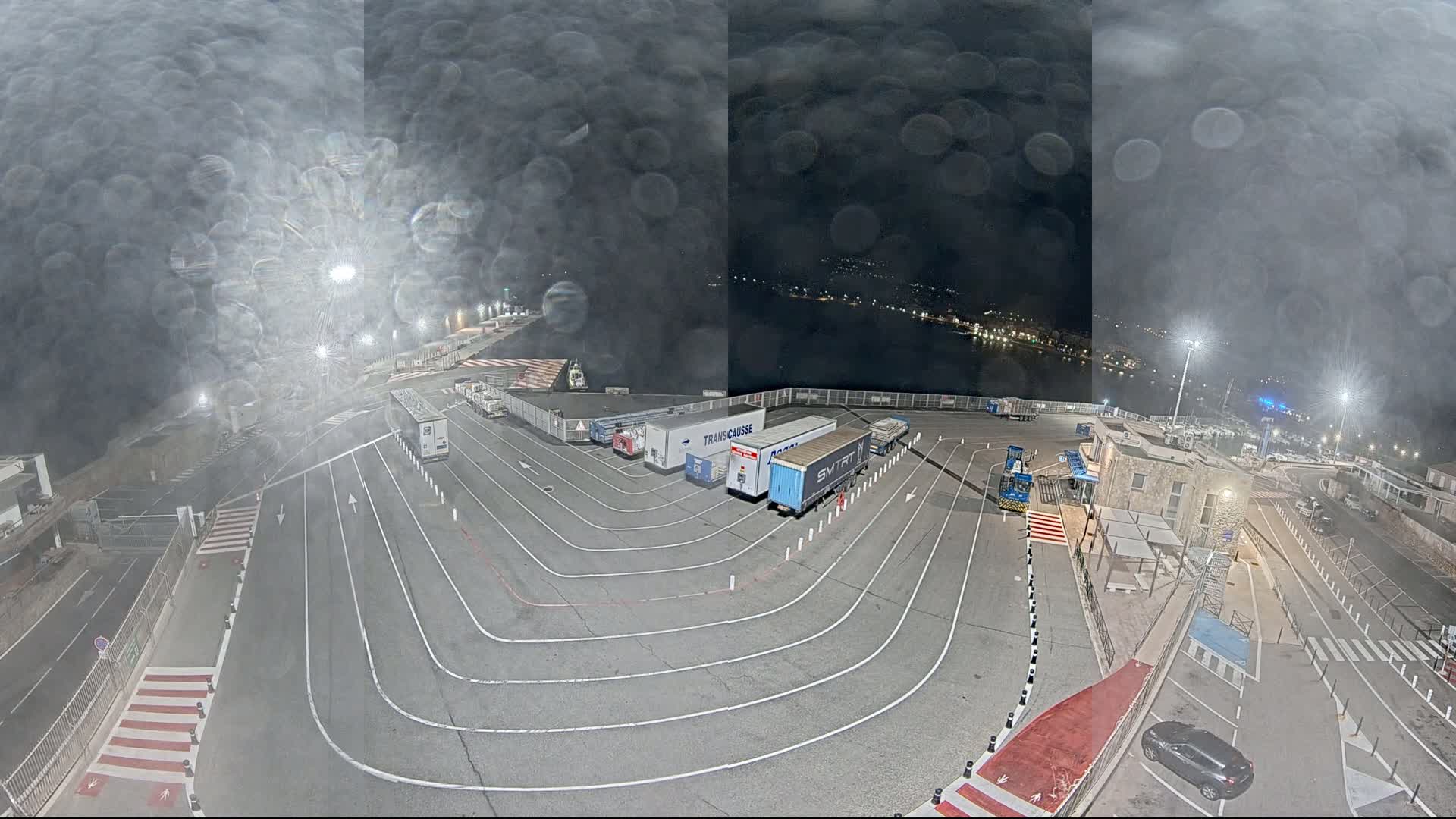 A nighttime aerial view of a port area with several trucks and trailers parked in designated lanes, under a hazy, rain-streaked sky.