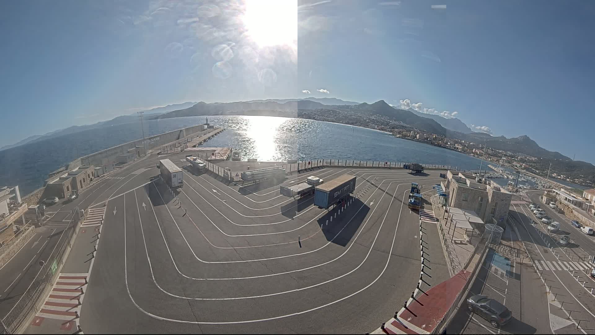 A sunny day at a harbor shows a large, curved roadway lined with trucks and cars, overlooking a calm body of water and distant mountains.