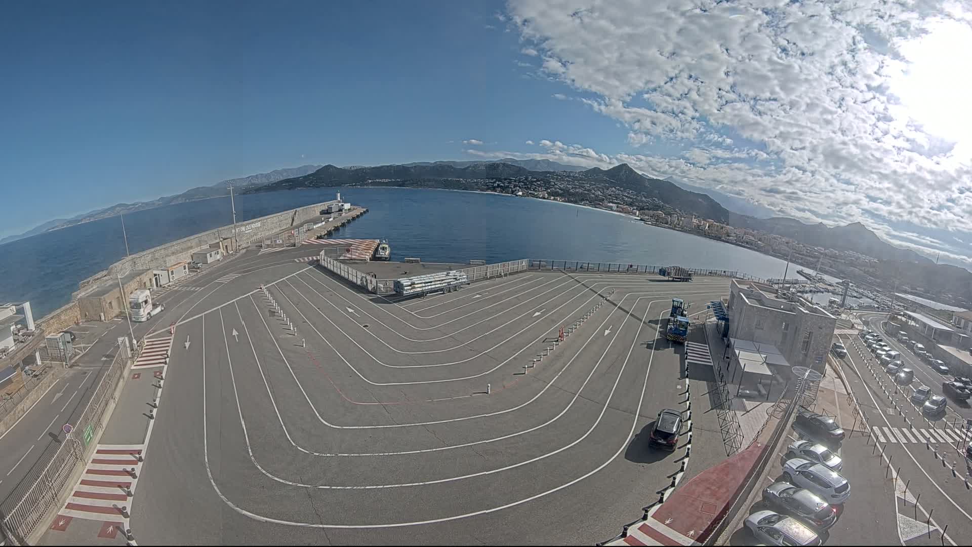 A mostly sunny day overlooks a harbor with a large, empty, marked parking lot and several cars parked nearby, mountains in the background.