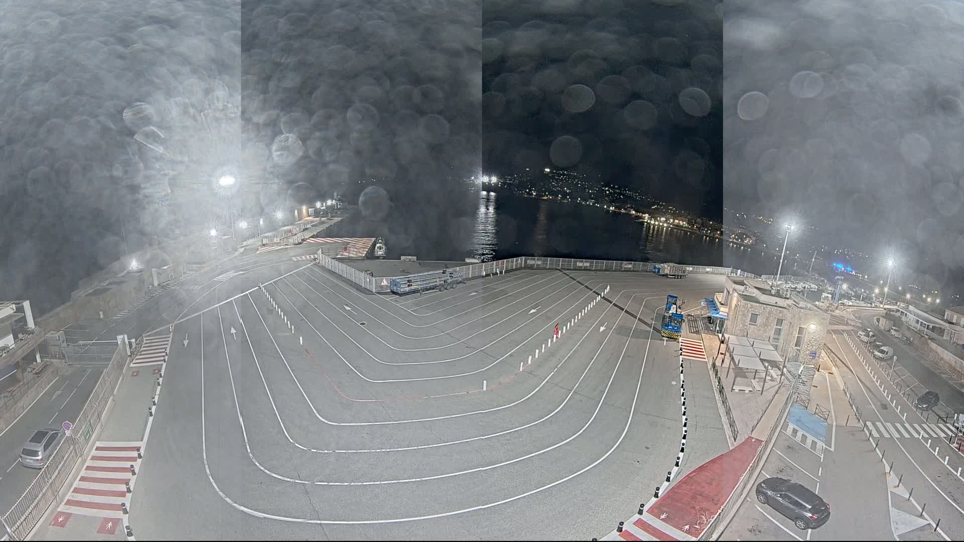 A nighttime view of a ferry terminal with a large, empty vehicle staging area, illuminated by lights, under a hazy, possibly rainy sky.