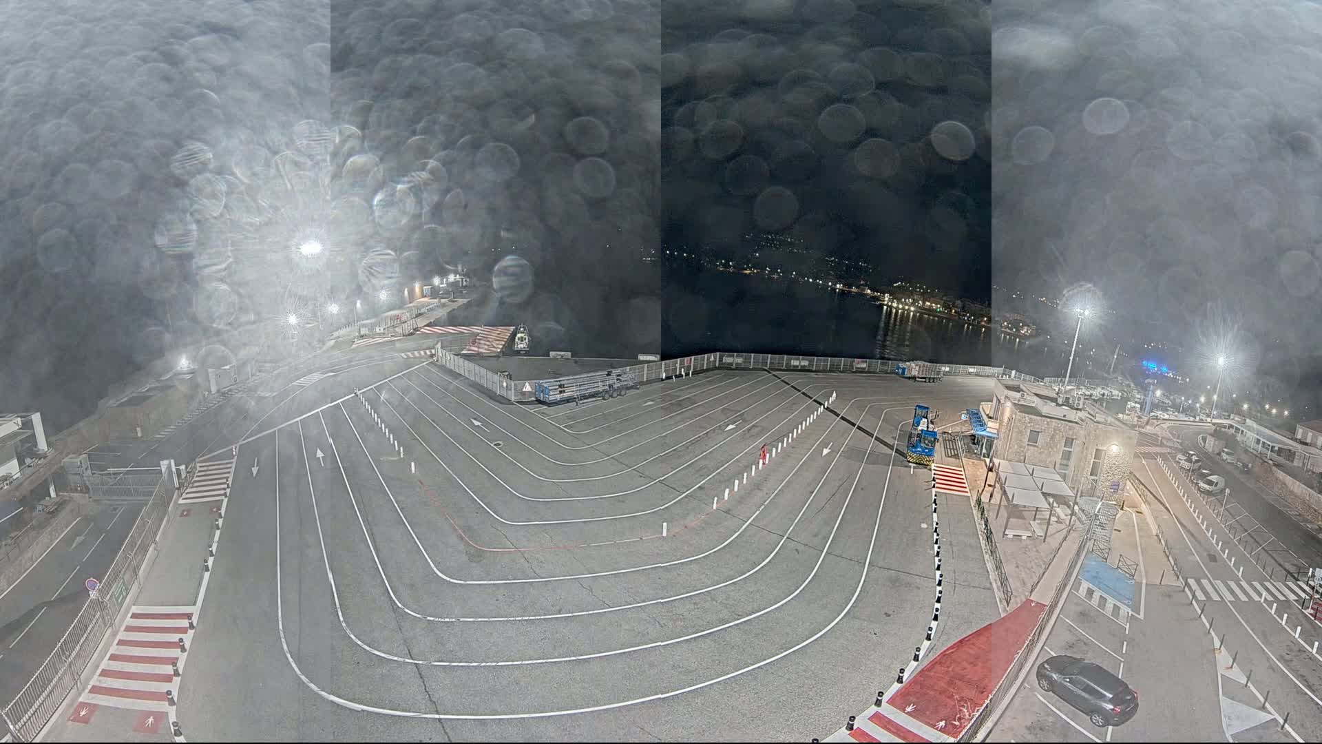 A nighttime view of an empty ferry terminal with rain obscuring the view, showing a large paved area with marked lanes and a few vehicles.