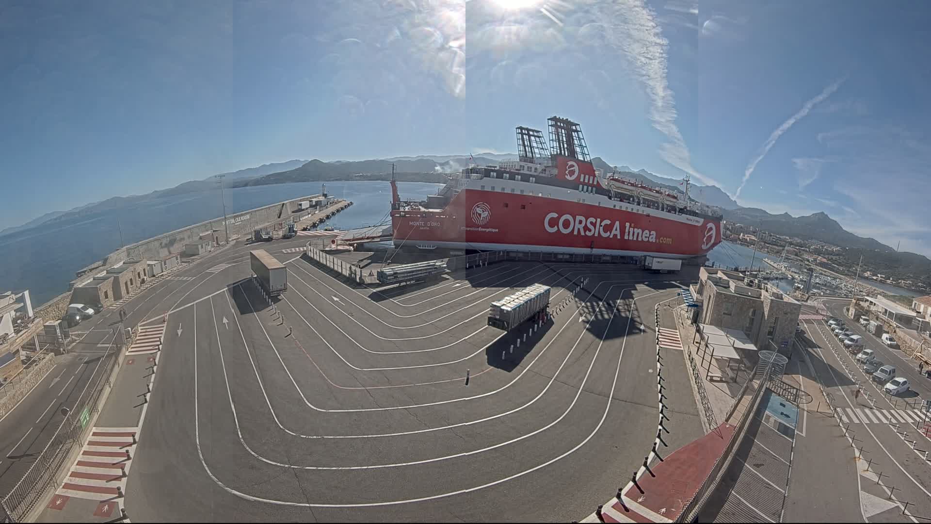 A large red ferry is docked at a port on a sunny day, with trucks and cars in the surrounding parking area and mountains visible in the distance.