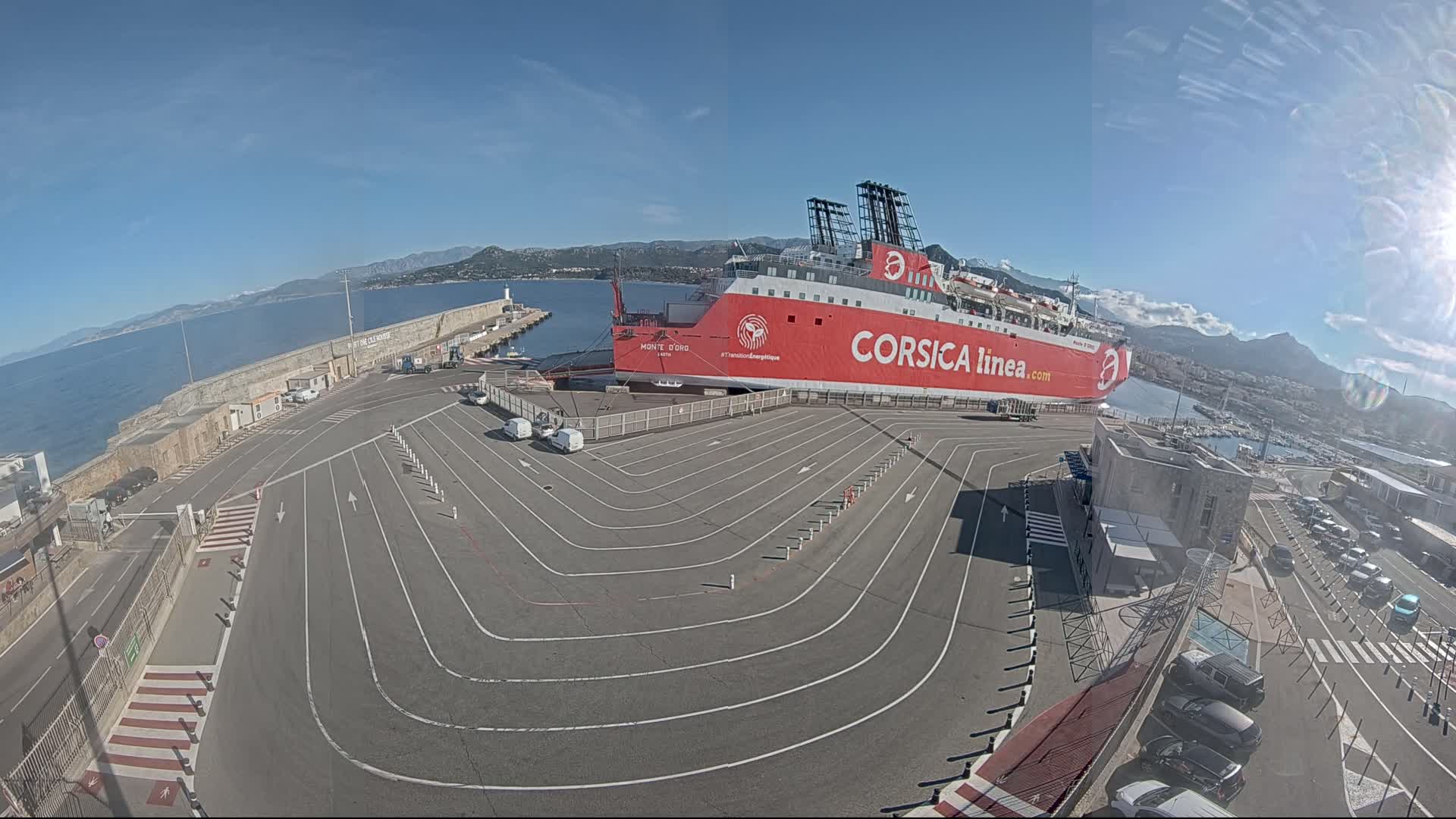 A large red ferry is docked at a port on a sunny day, with mountains visible in the background and a parking lot with lane markings in the foreground.