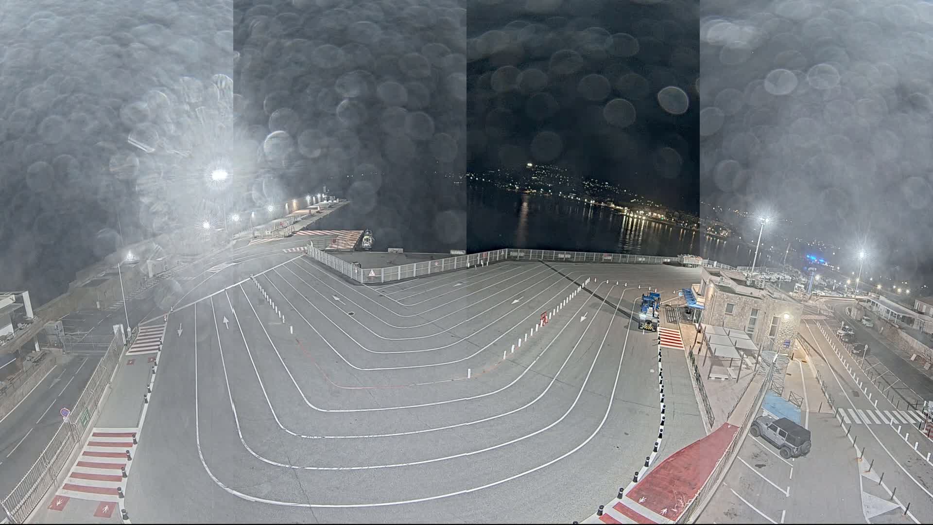A nighttime view of a port's vehicle staging area with a body of water in the background, viewed through a rain-covered lens.