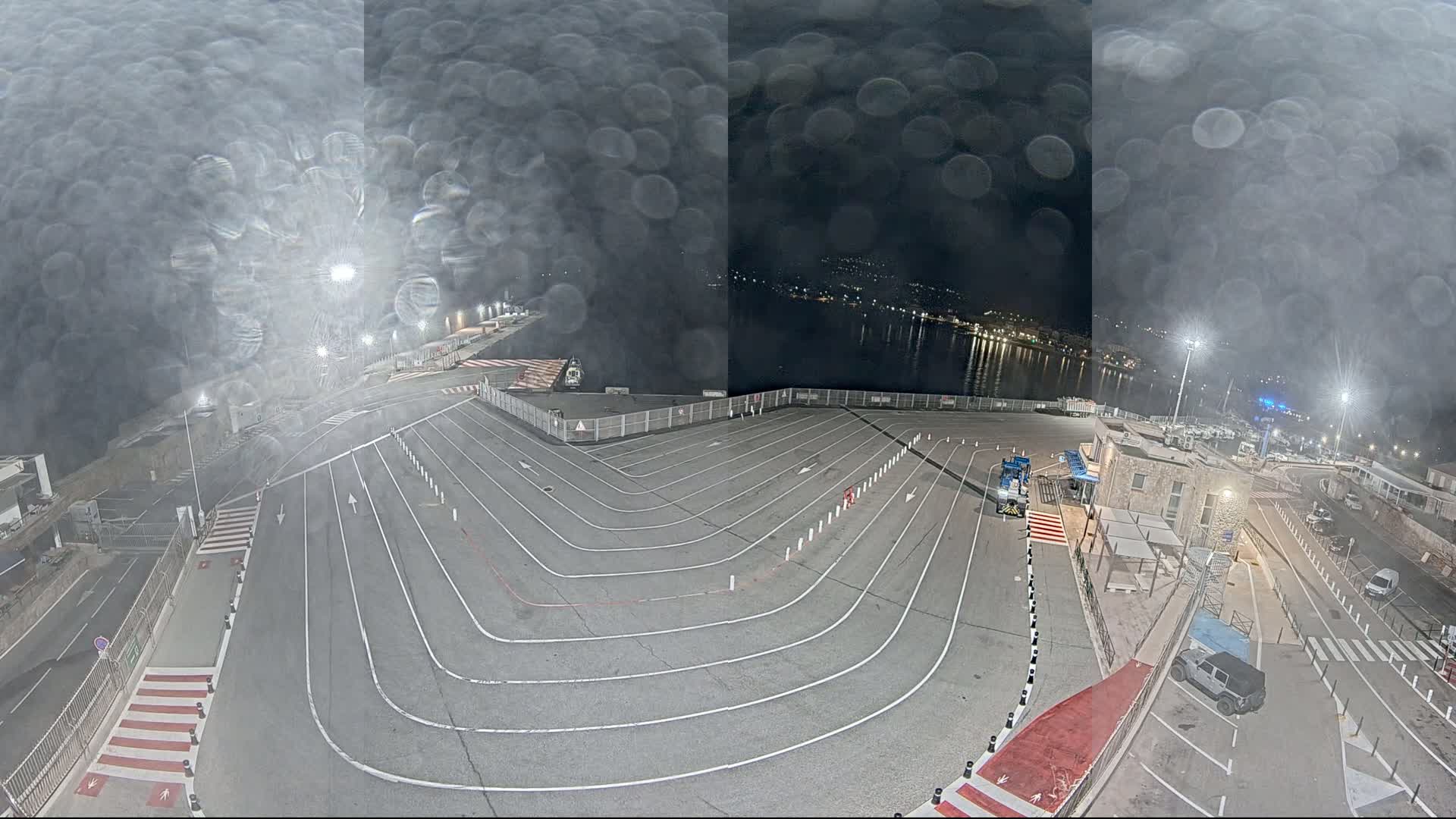 A nighttime view of a ferry terminal with a large, marked parking area, viewed through rain-streaked glass, shows a calm body of water and a town in the distance.