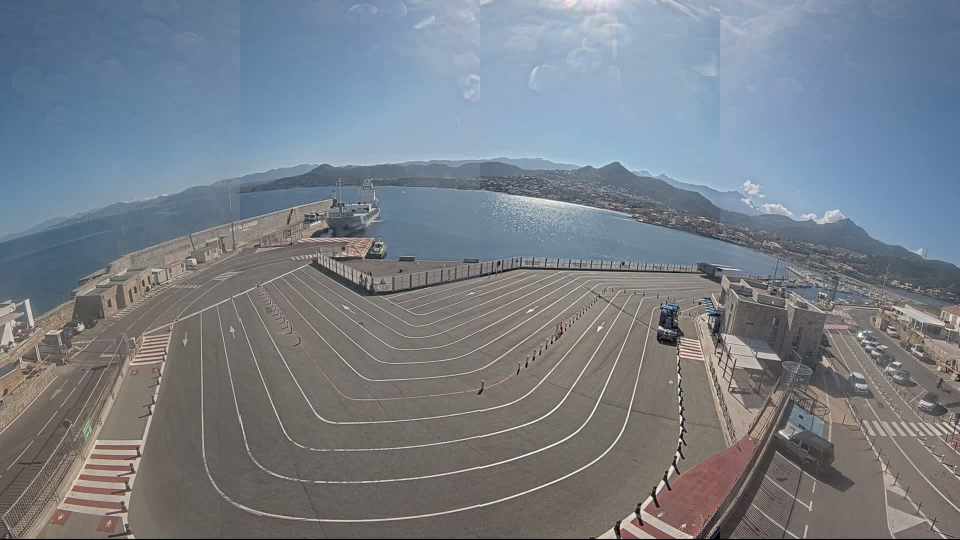 A large, empty paved area with marked lanes curves down to a harbor where a ship is docked, all under a sunny sky with mountains in the background.