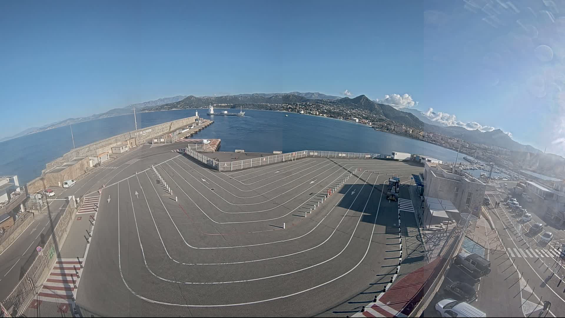 A large, empty vehicle staging area at a port curves around a harbor, with a calm sea and distant mountains under a clear, sunny sky.