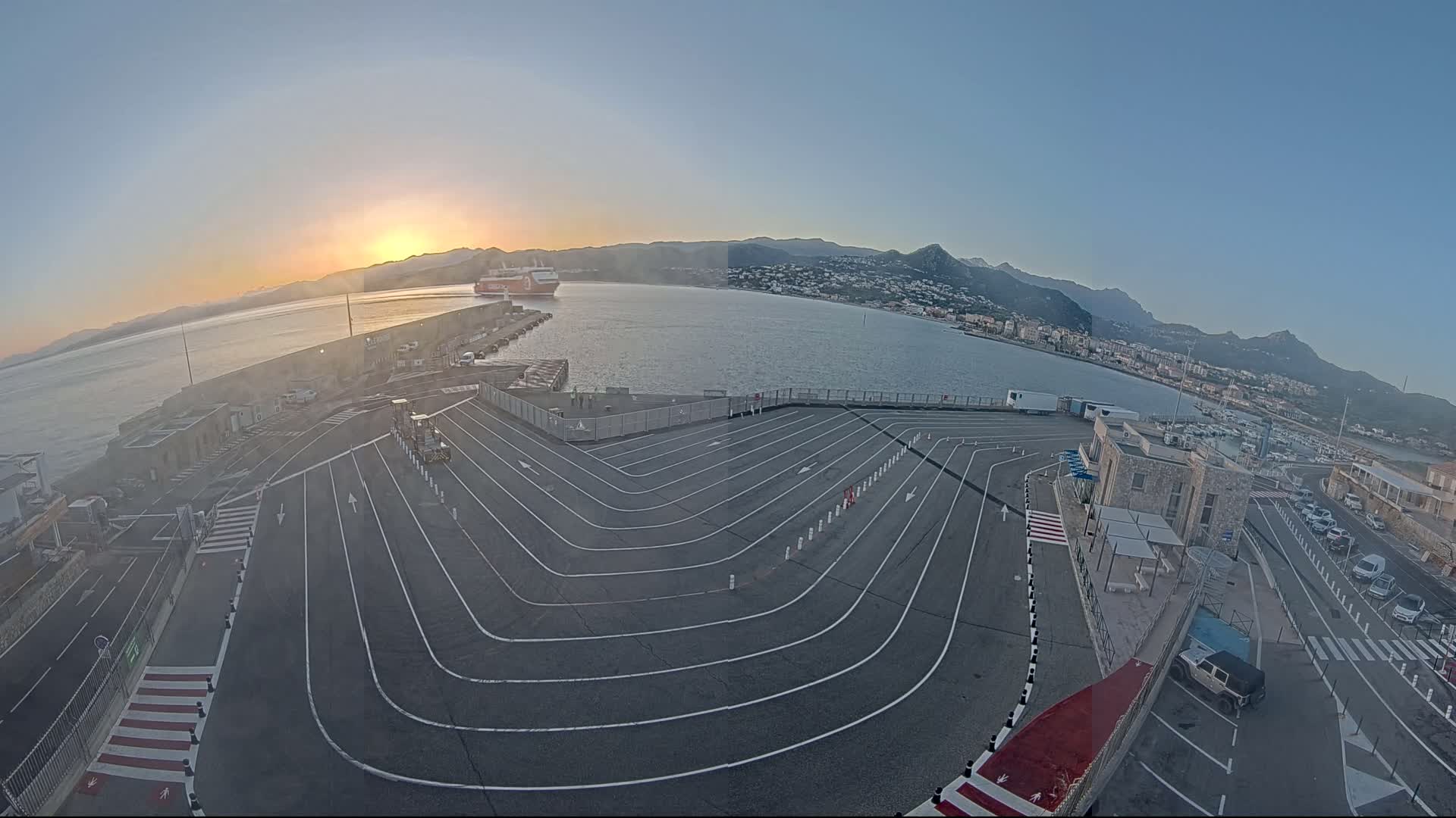 A large, curved vehicle staging area at a port, with a ferry visible in the distance, under a clear sunrise sky.