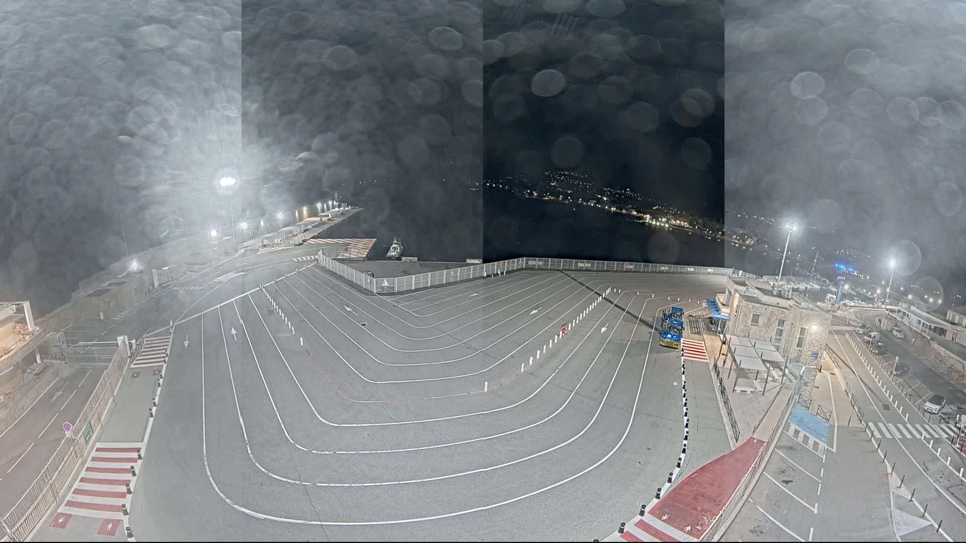 A nighttime view of a ferry terminal with a large, empty vehicle staging area, lit by floodlights, under a misty or rainy sky.