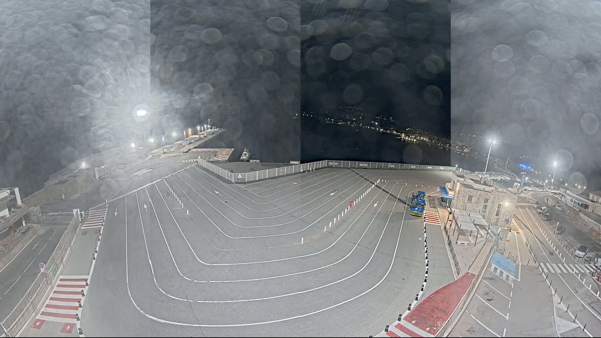 A nighttime view of a port's vehicle staging area with a rain-streaked lens shows a large, empty paved lot with marked lanes and a few vehicles near a small building.