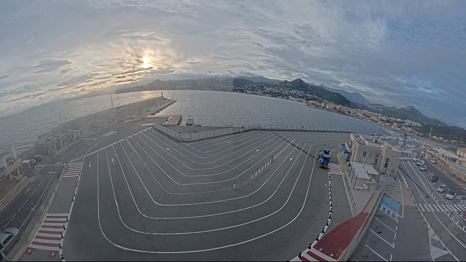 A ferry terminal with a large, winding roadway is situated by a calm body of water under a partly cloudy sky with the sun setting behind distant mountains.