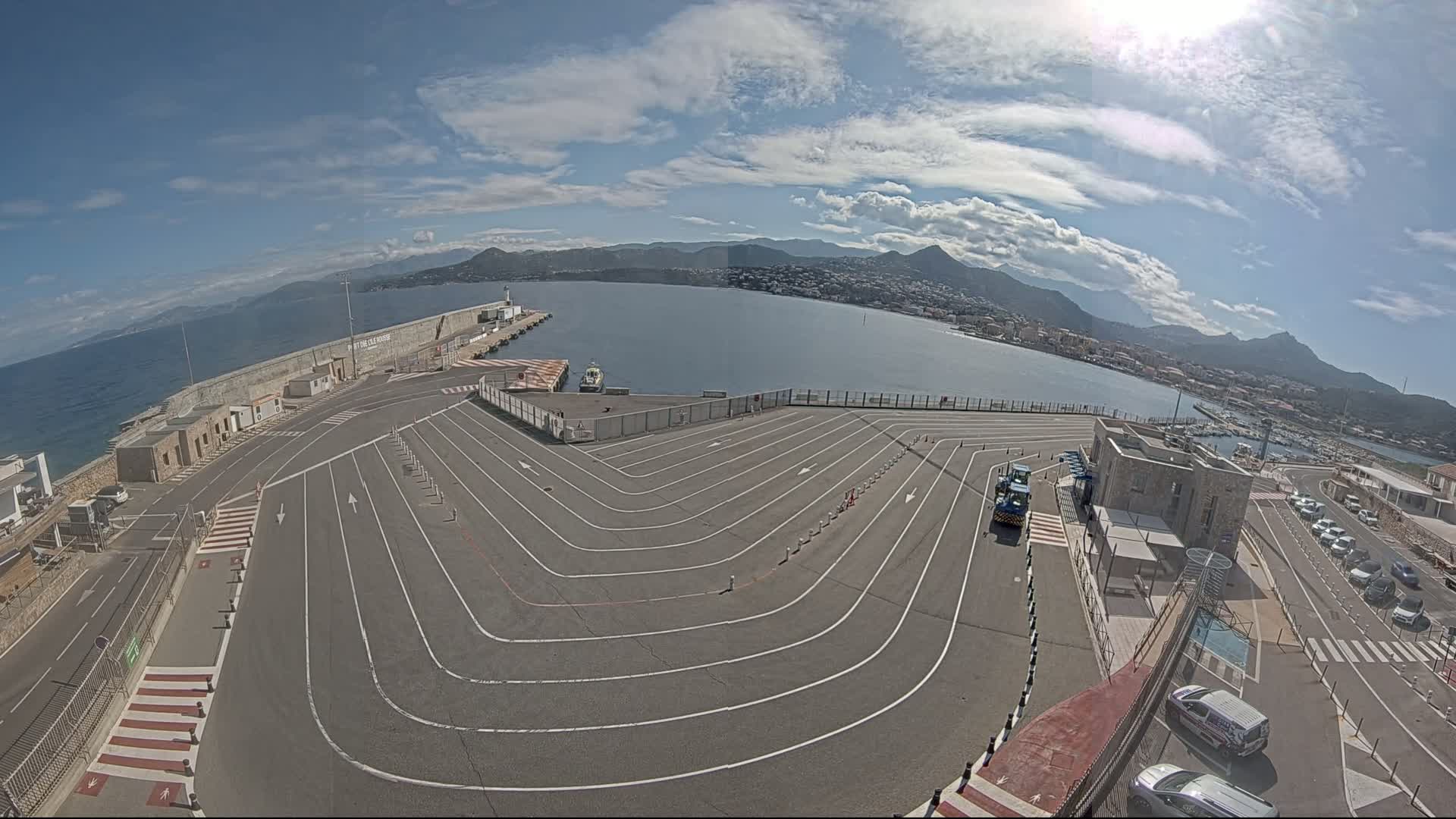 A large, empty paved area with marked lanes curves toward a harbor, overlooking a calm sea and distant mountains under a mostly sunny sky with some clouds.