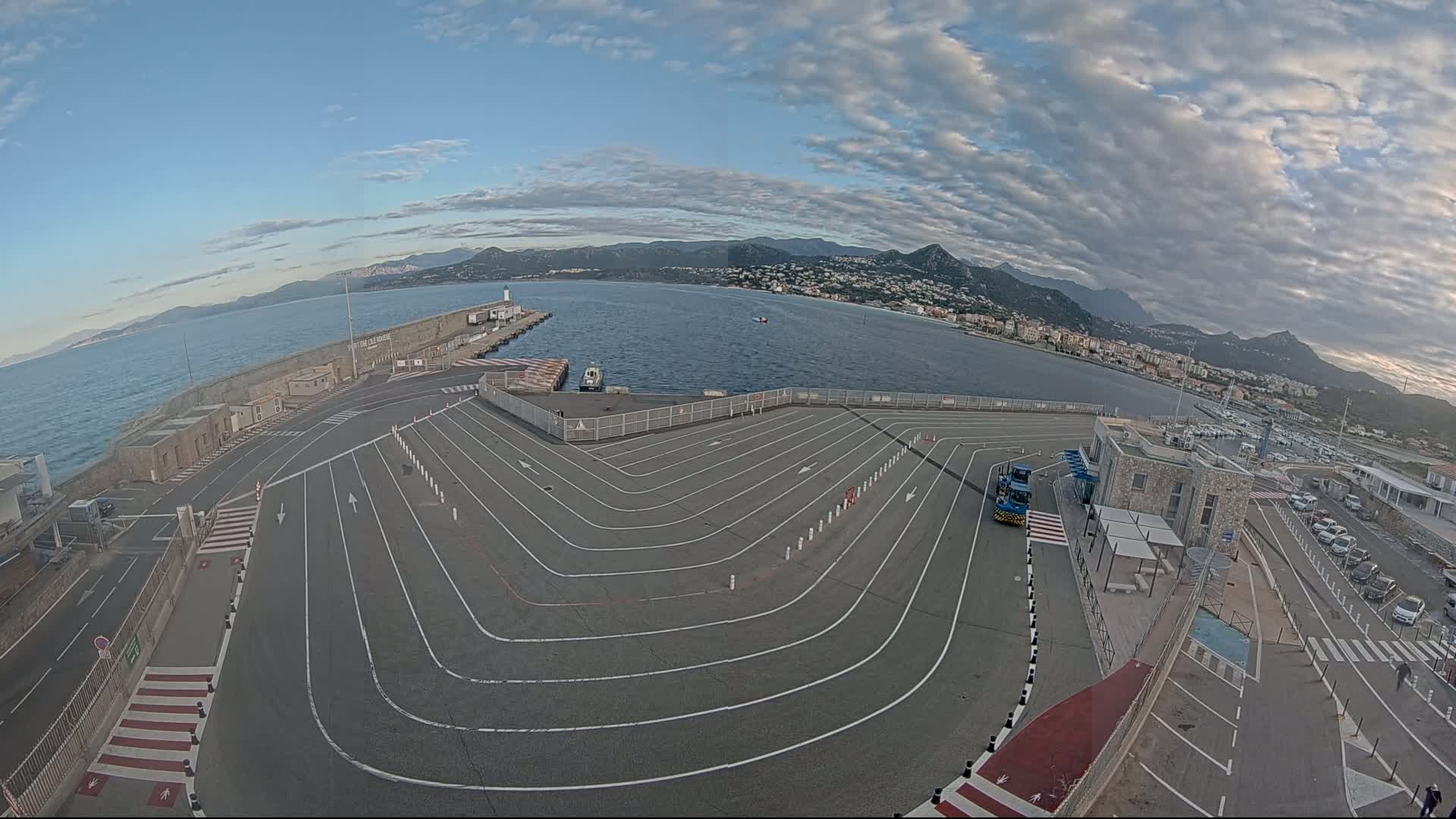 An aerial view of a ferry terminal with a large, marked parking area, overlooking a calm body of water under a partly cloudy sky, with mountains visible in the distance.