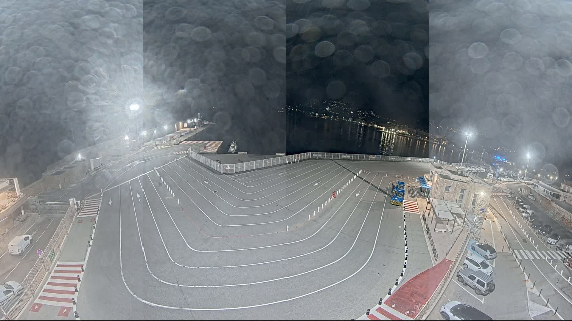 An empty, marked vehicle staging area at a port at night, under a misty sky, overlooks a dark body of water with city lights in the distance.