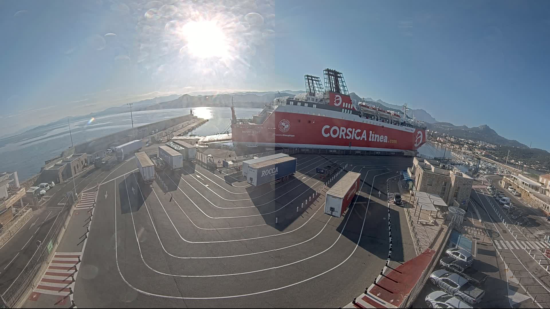 A large red ferry is docked at a port on a sunny day, surrounded by trucks and cars in a paved area with mountains visible in the distance.