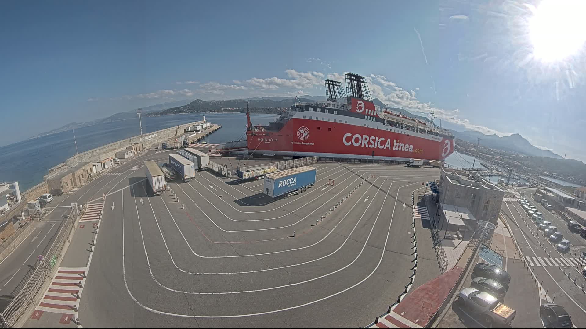 A large red ferry is docked at a port on a sunny day, with numerous trucks and cars parked in designated lanes nearby.