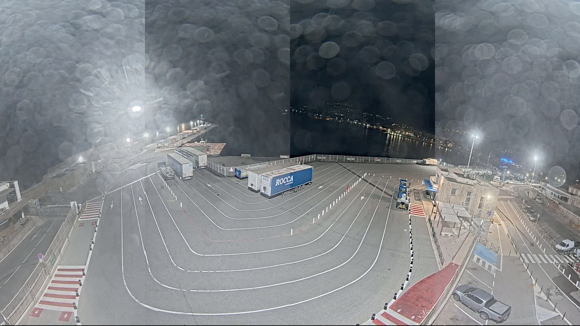 A nighttime aerial view of a port area shows several large trucks parked in designated lanes, with a body of water visible in the background under a slightly hazy sky.