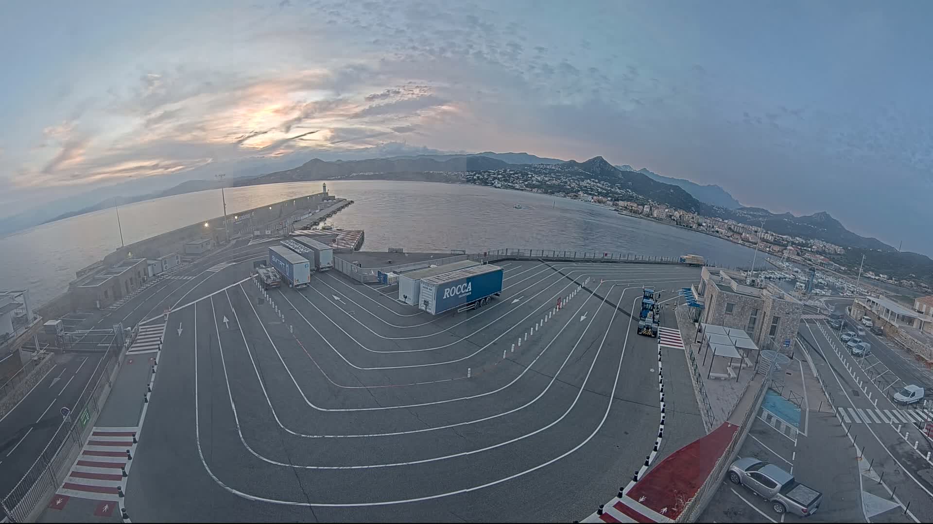 A port at sunrise, with several semi-trailer trucks parked in a large, marked lot, overlooks calm water and distant mountains.