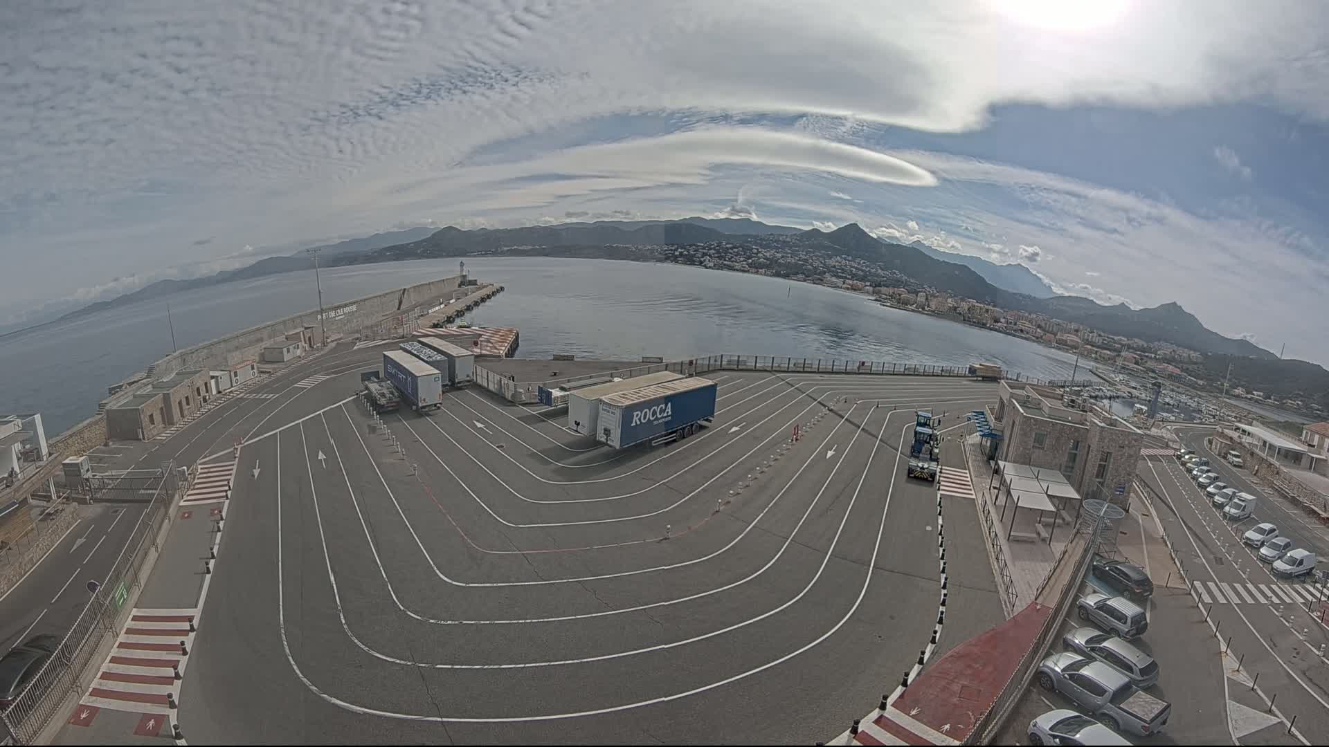 An aerial view of a port with several semi-trailer trucks parked in a designated area, under a partly cloudy sky, overlooking a calm body of water and mountains in the distance.