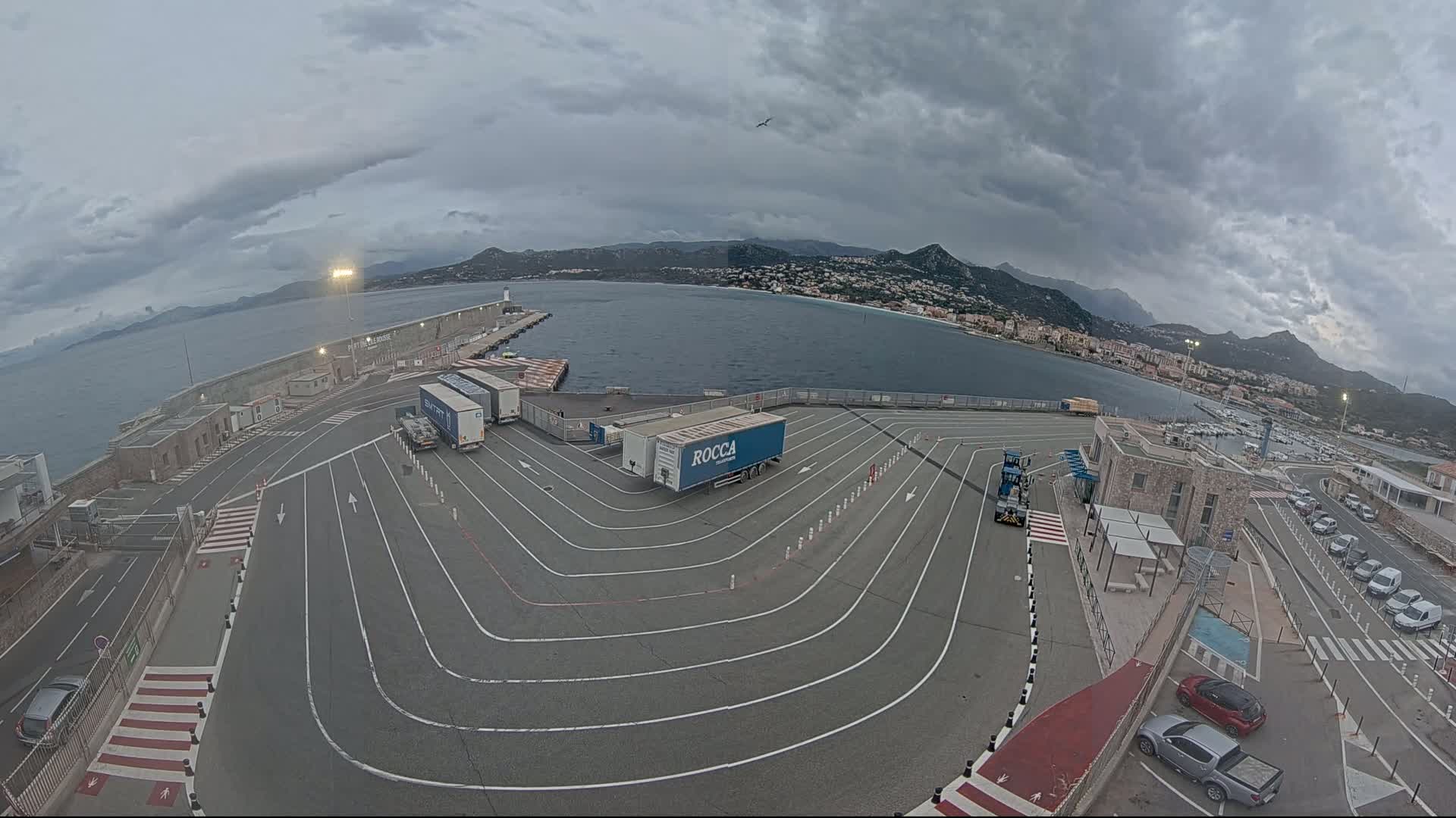 A ferry terminal with several trucks parked in a curving roadway, overlooking a calm body of water under a cloudy sky.