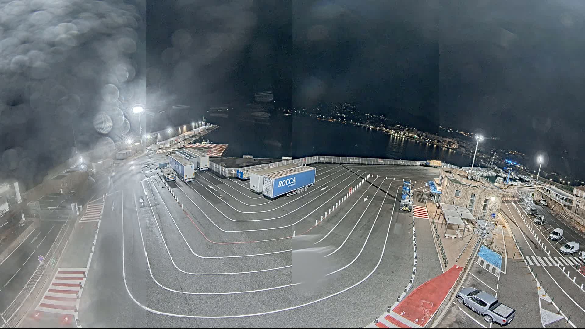A nighttime view of a port with several trucks parked in a designated area under a dark, possibly rainy sky.