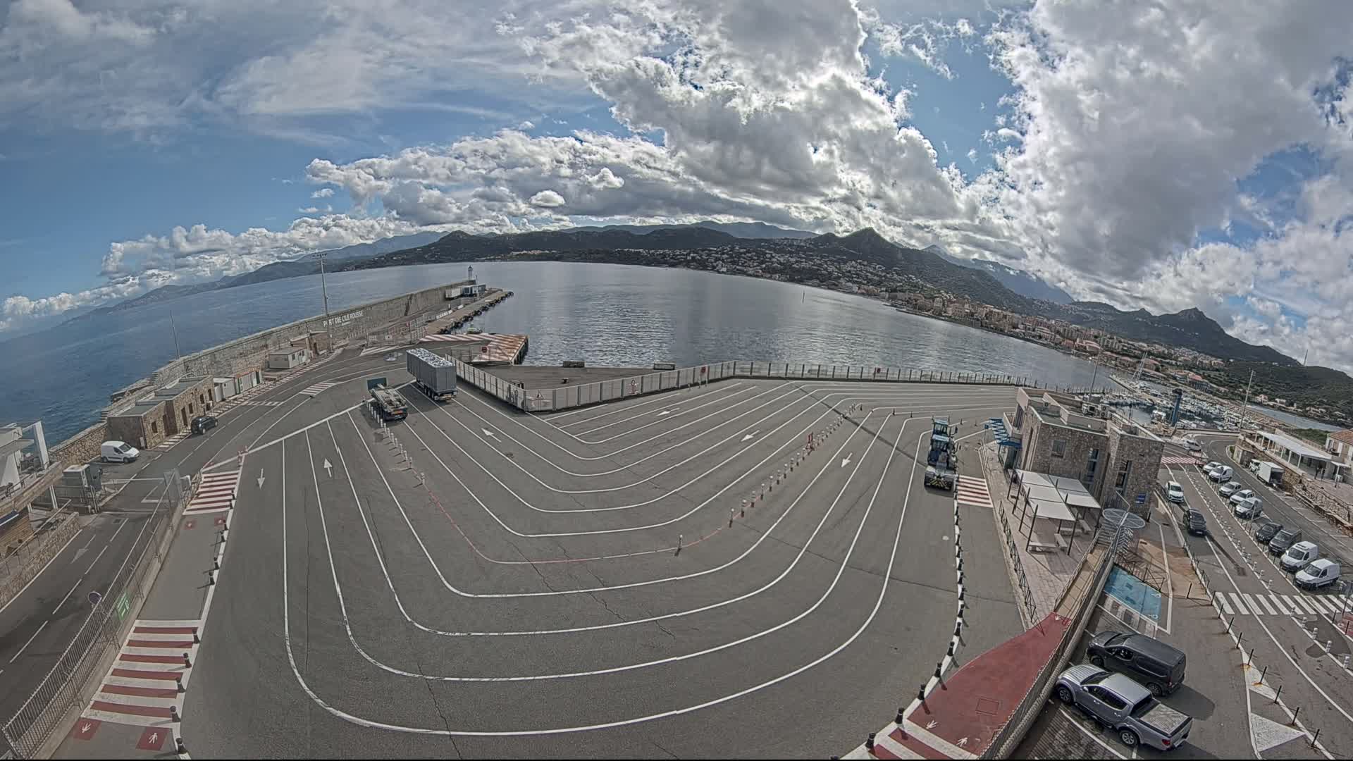 A partly cloudy day overlooks a harbor with a large, curved vehicle staging area, a small building, and several cars parked nearby.