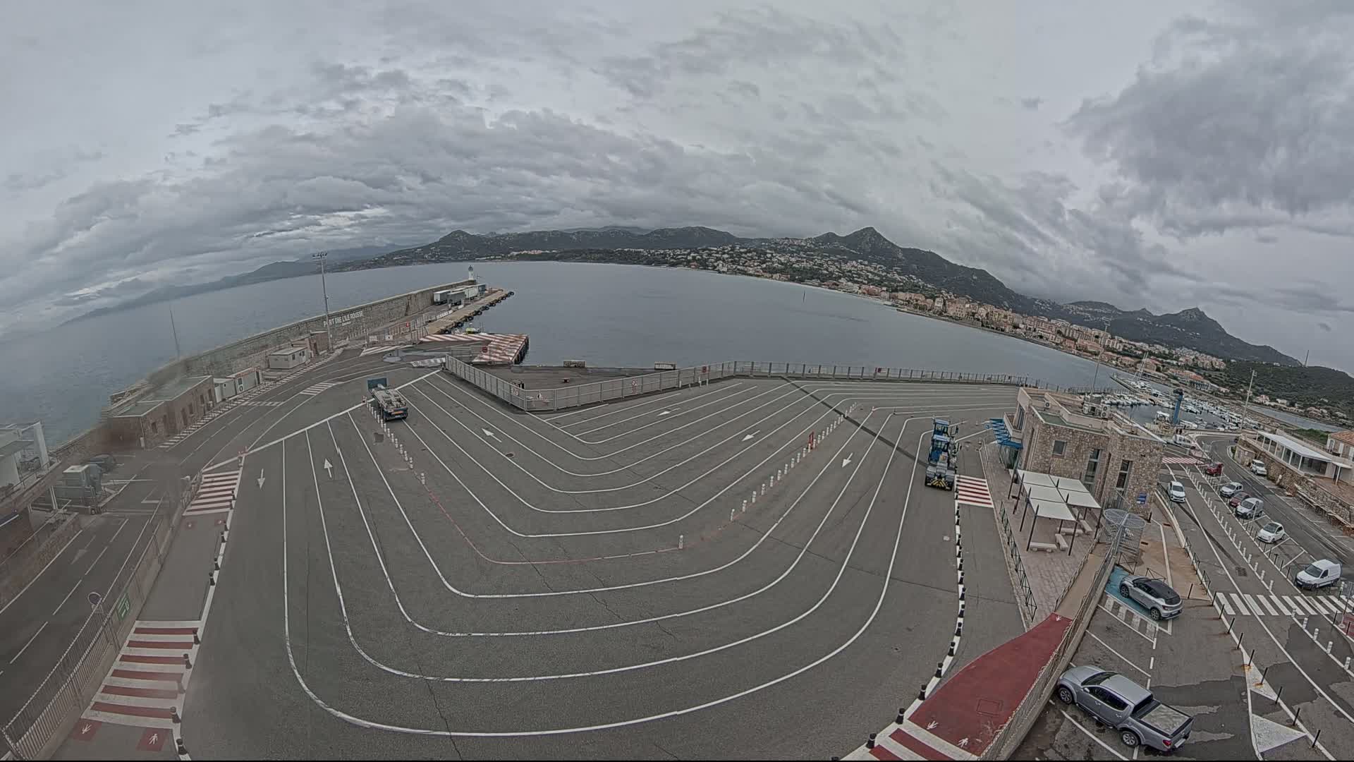 An overcast day reveals a harbor's ferry terminal with a large, marked vehicle queuing area, overlooking a calm body of water and distant hills.