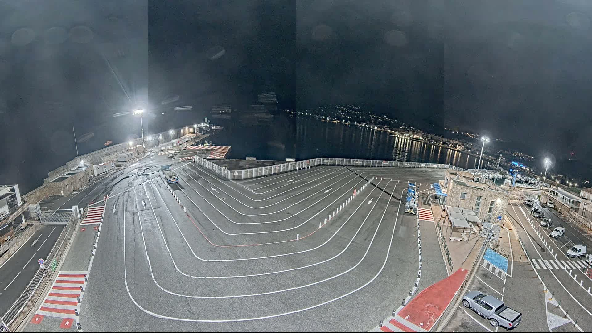 An empty, brightly lit ferry terminal at night, with a calm body of water and a town visible in the distance under a dark, cloudy sky.