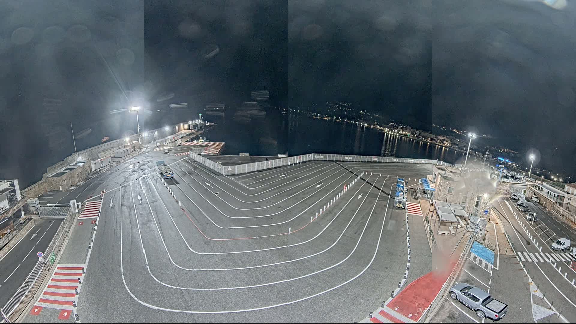 A nighttime aerial view shows a ferry terminal's empty vehicle lanes curving toward a waterfront under a dark, cloudy sky.