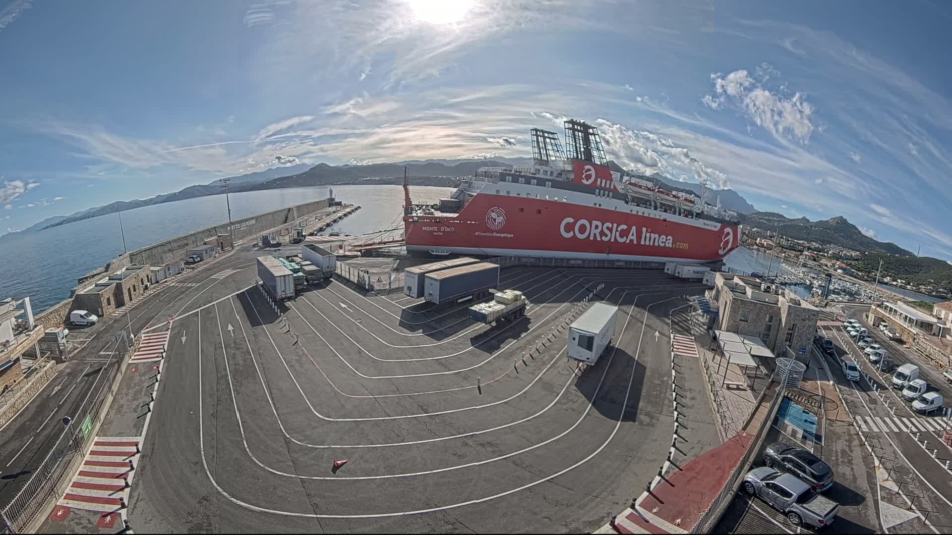 A large red and white ferry is docked at a port on a sunny day, with several trucks and cars parked nearby, and mountains visible in the distance.