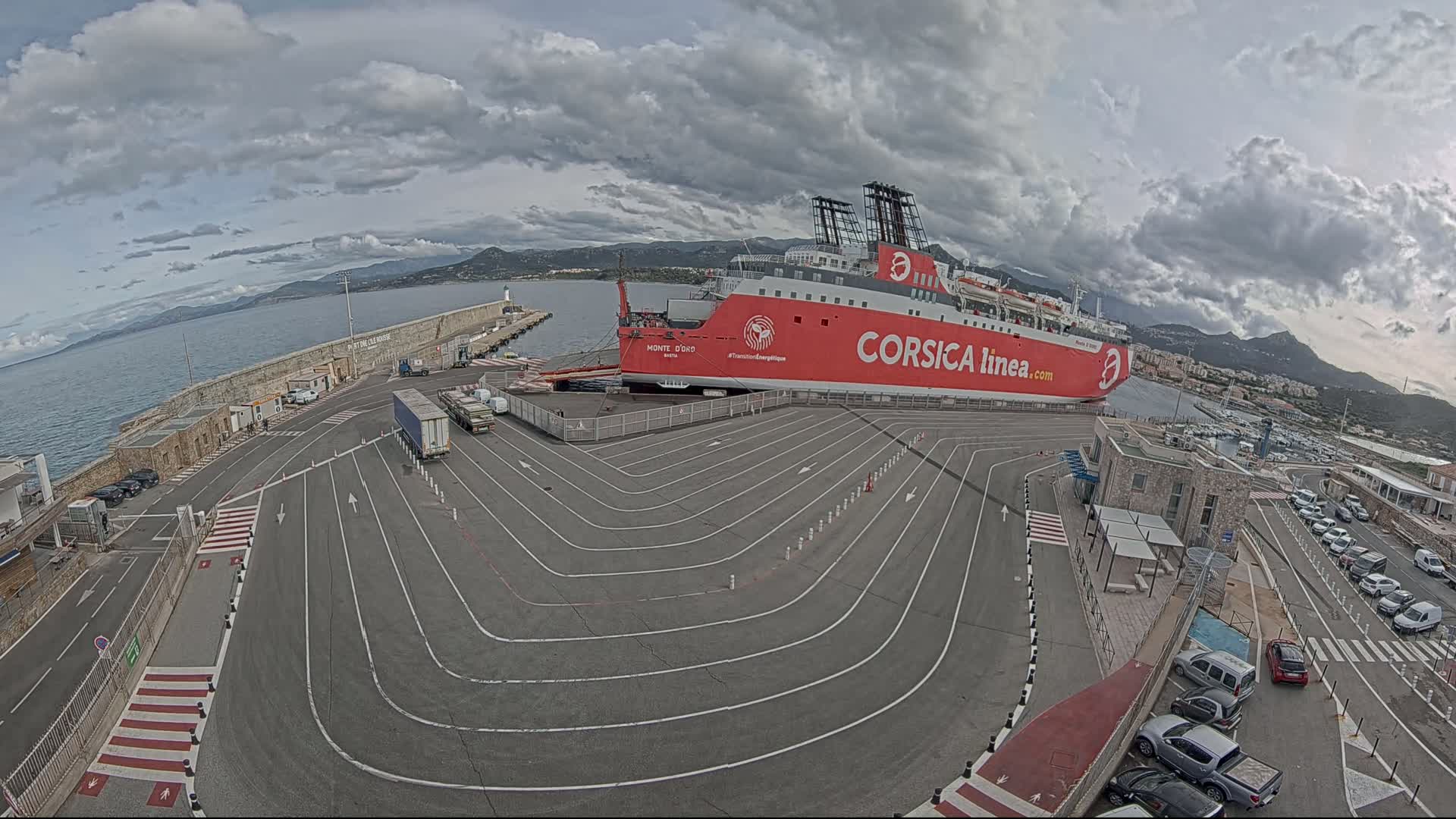 A large red ferry is docked at a port under a cloudy sky, with marked lanes for vehicles in the foreground and cars parked nearby.
