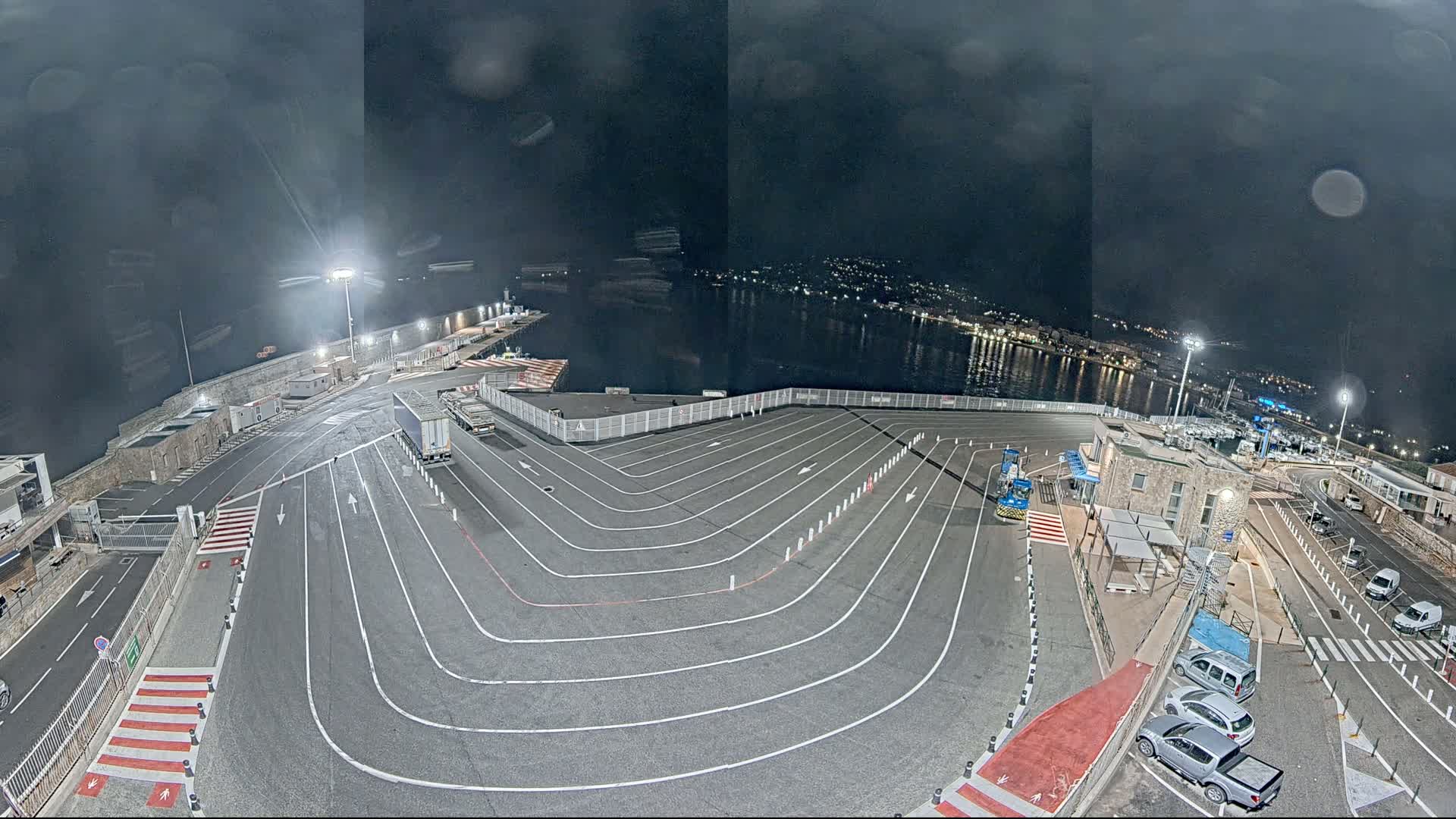 A nighttime aerial view of a ferry terminal with marked lanes for vehicles, a few cars parked nearby, and a body of water with city lights in the background under a dark, cloudy sky.