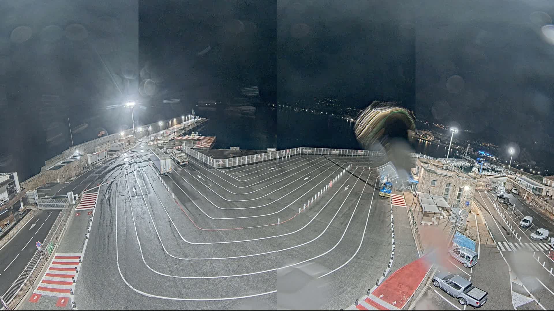A wet, nighttime aerial view of a harbor's ferry terminal with marked vehicle lanes, several parked cars and trucks, and a body of water visible in the background.
