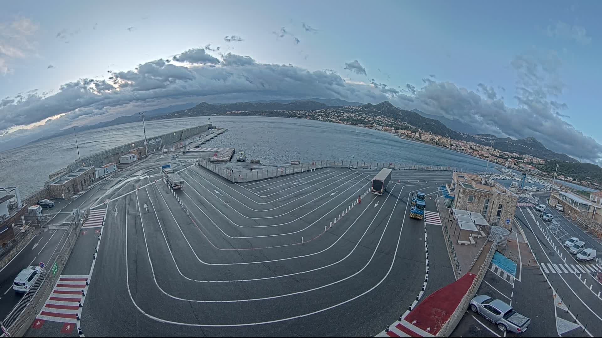 An aerial view of a ferry terminal with several vehicles parked, under a cloudy sky overlooking a calm body of water and distant mountains.