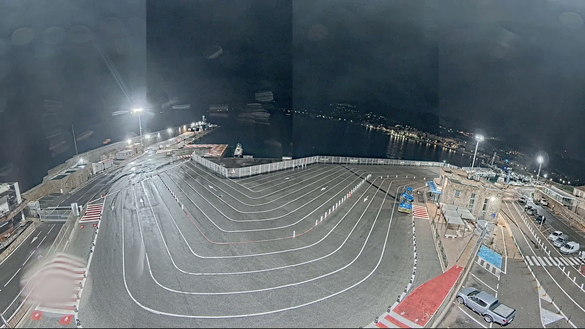 An empty, nighttime ferry terminal with a large, curved roadway marked with white lines, illuminated by artificial lights, overlooks a calm body of water.