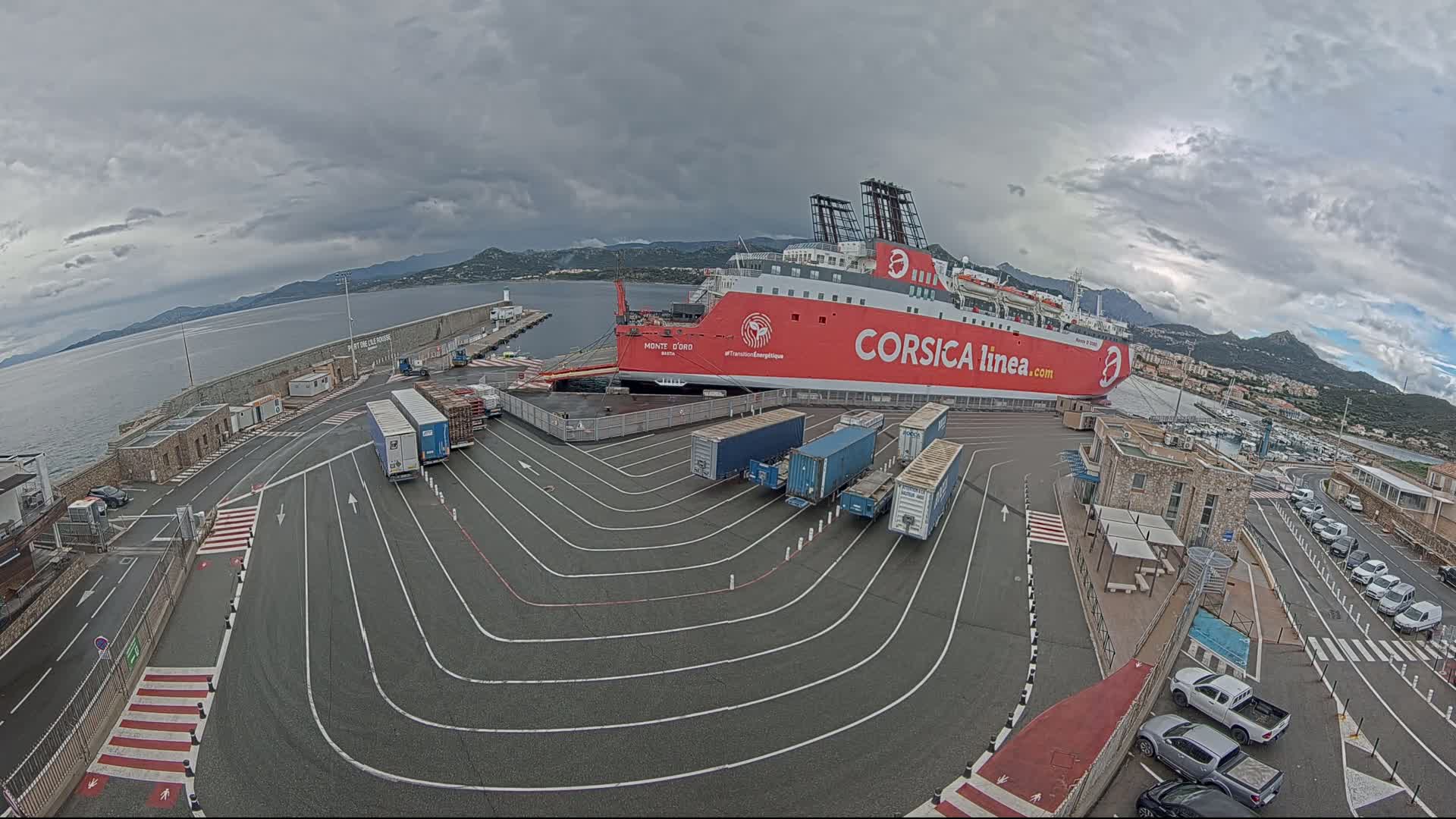 A large red and white ferry is docked at a port under an overcast sky, with several semi-trailer trucks parked nearby.