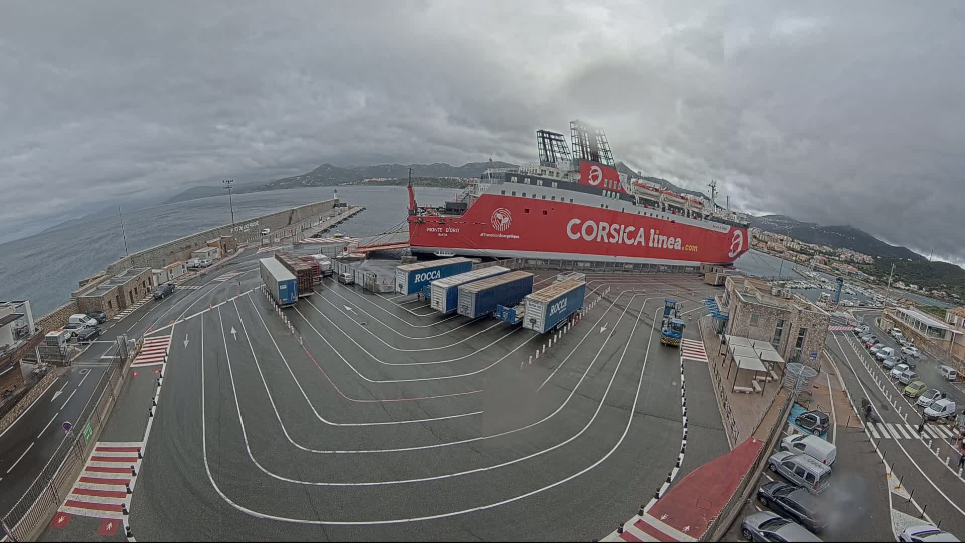 A large red ferry is docked at a port under an overcast sky, with several semi-trailer trucks parked nearby.