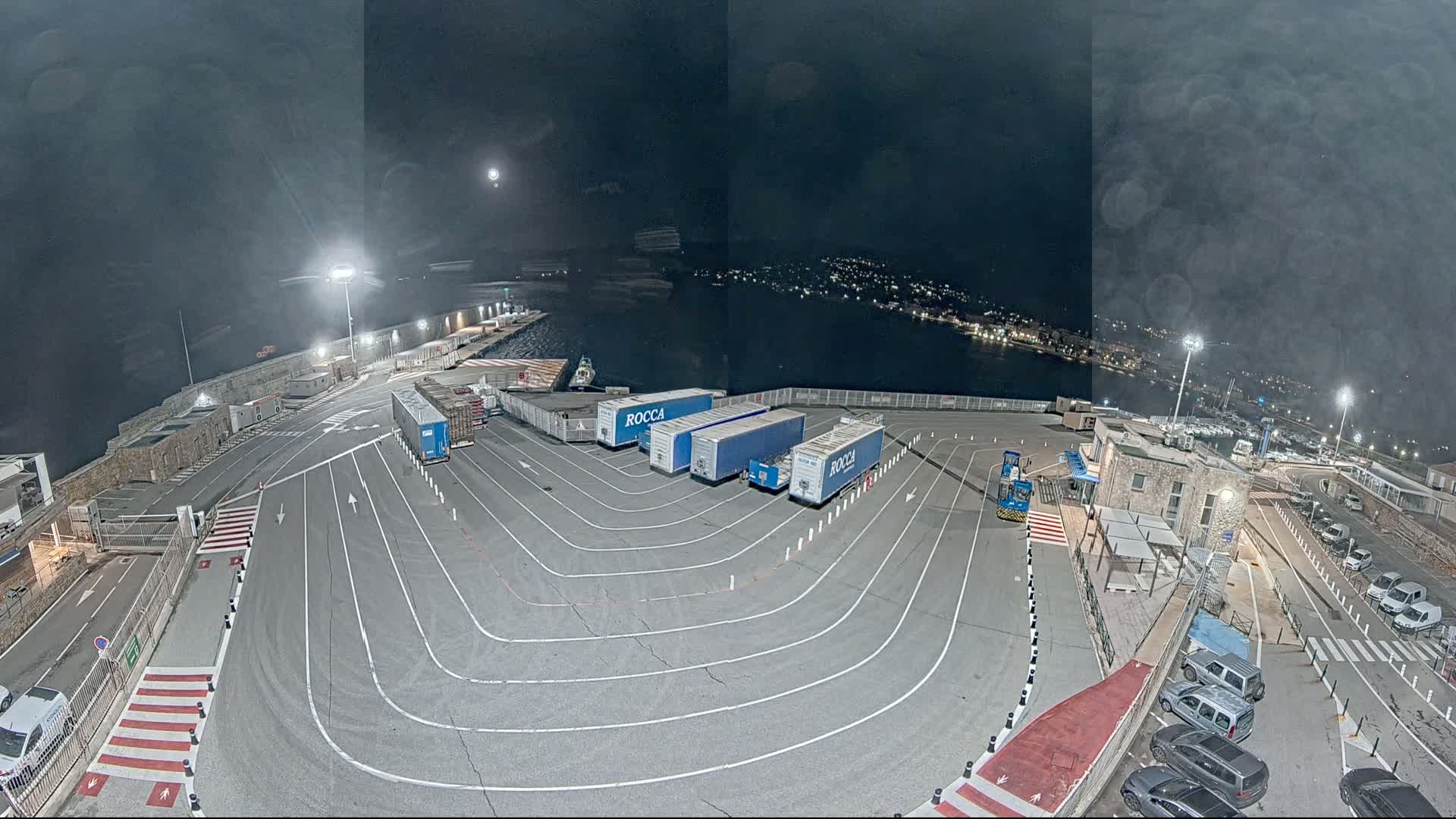 At night, a ferry terminal with several semi-trailer trucks is seen parked in designated areas, under partly cloudy skies.