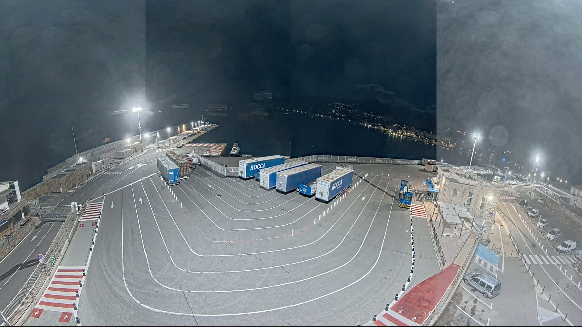 At night, a port area shows several blue semi-trailer trucks parked in designated lanes, with a few cars in a nearby lot, under a dark, mostly cloudy sky.