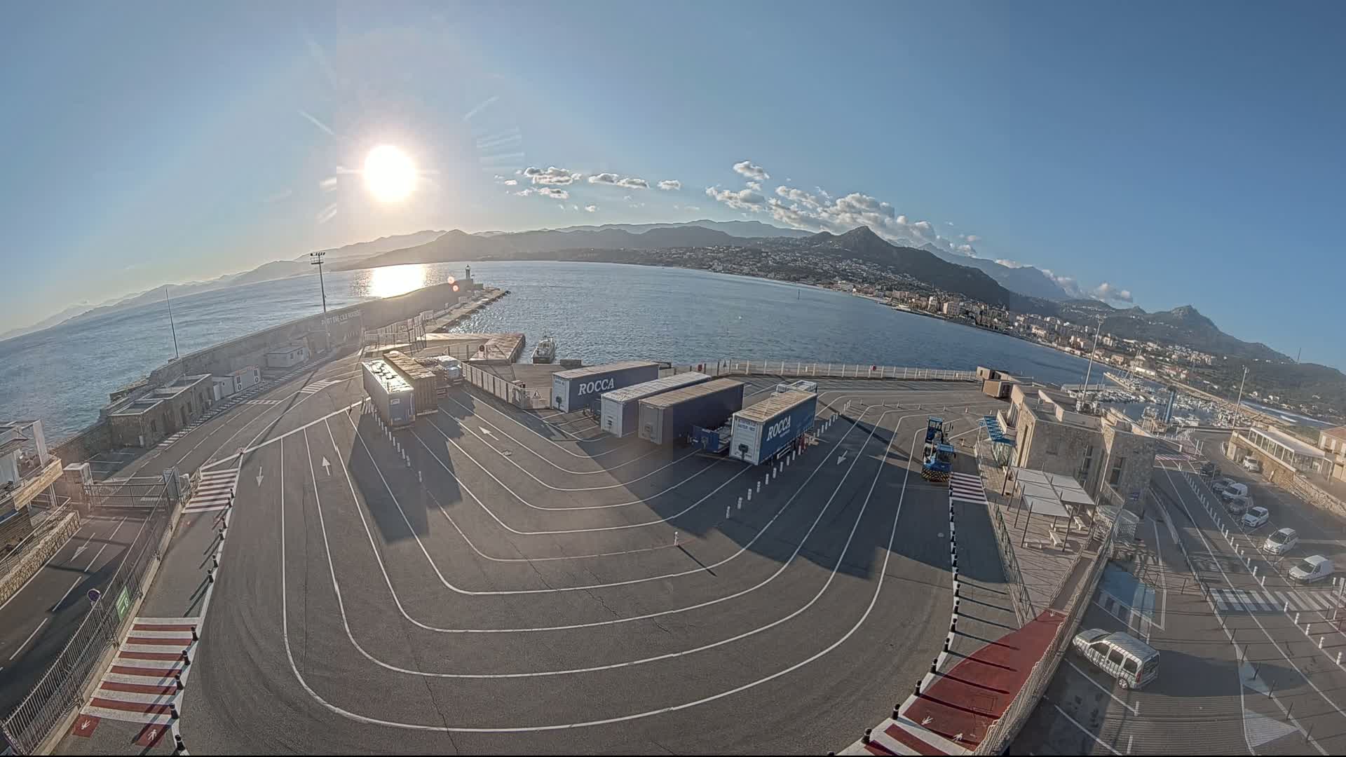 An aerial view of a port area on a sunny day, showing several large trucks parked in a designated area with a body of water and mountains visible in the background.